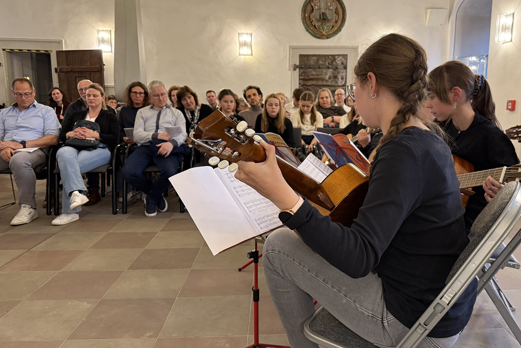 Zwei Mädchen mit Gitarre vor einem größeren Publikum auf Stühlen