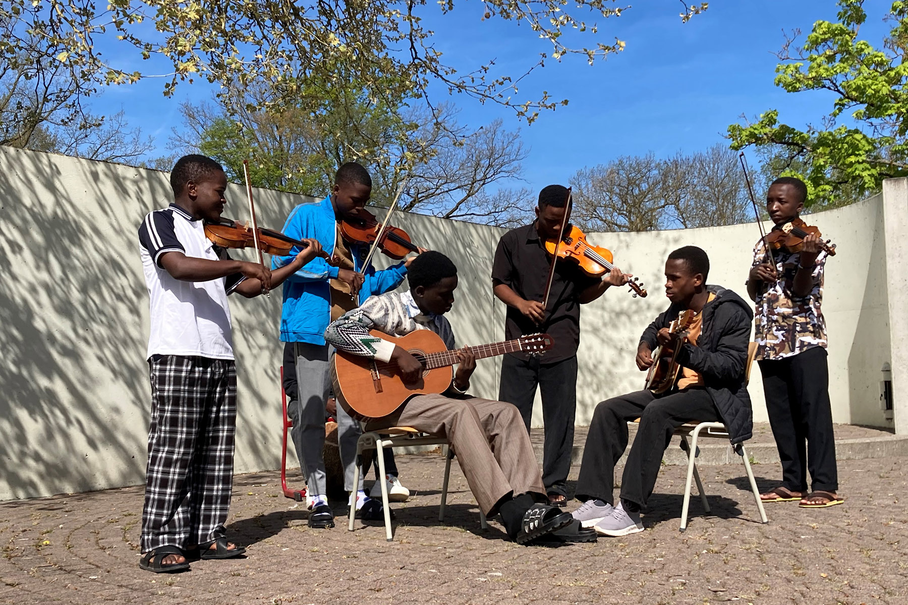 Sechs Schüler aus Tansania spielen vor einer Mauer auf Geigen und Gitarren