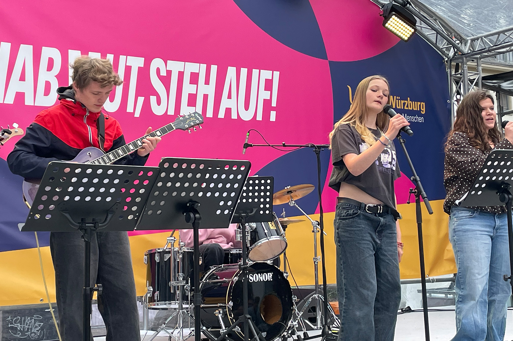 Gitarrist und zwei Sängerinnen der Schulband auf der Bühne vor einem bunten Banner