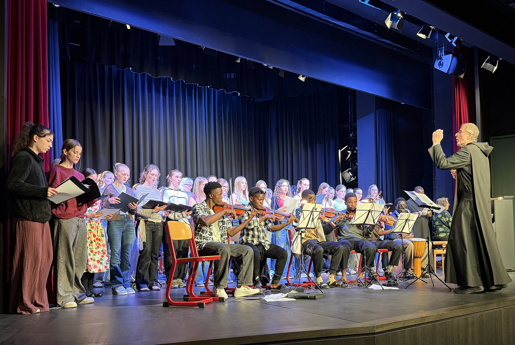 Sechs schwarze Schüler mit Geigen und Gitarren auf der Bühne, hinter ihn deutsche Schülerinnen und Schüler