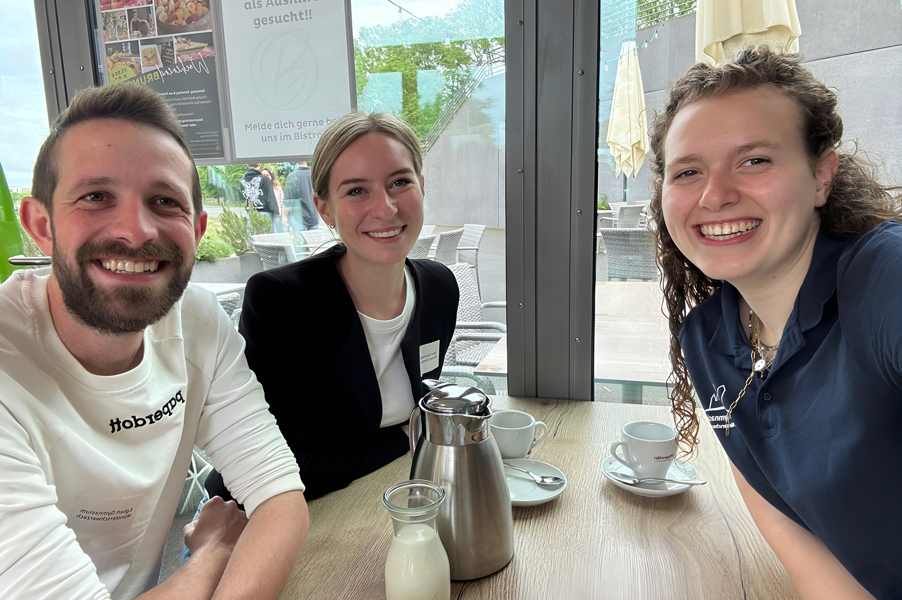 Mann mit Bart sitzt mit zwei Frauen an einem Tisch, auf dem eine Kaffeekanne und Tassen stehen