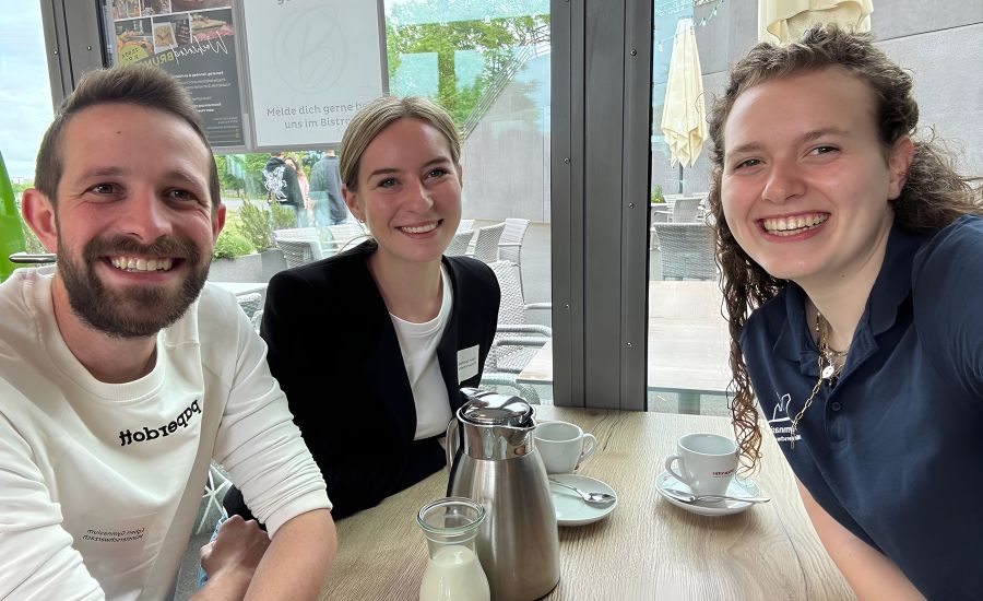 Ein Mann mit Bart und zwei Frauen sitzen an einem Tisch, auf dem eine Kaffeekanne und Tassen stehen