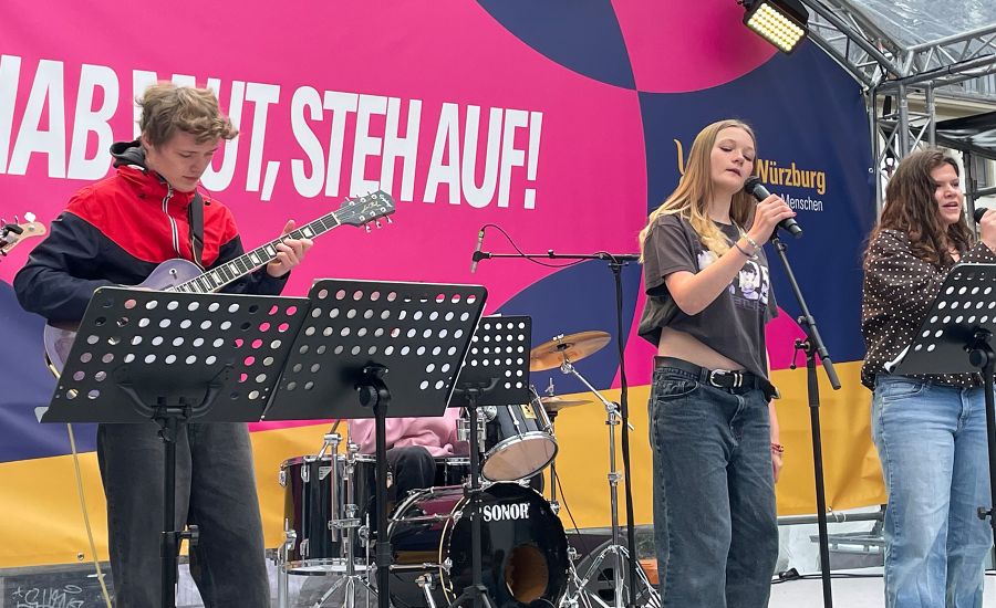 Gitarrist und zwei Sängerinnen der Schulband auf der Bühne vor einem bunten Banner